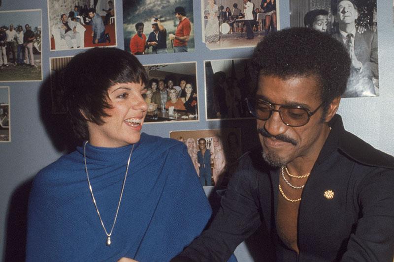 Sammy Davis smokes a cigarette while chatting with Liza Minelli in his dressing room, the walls of which are adorn with photos of Sammy throughout his career.