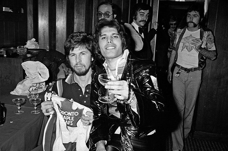 Freddie Mercury sits on his bandmate's lap while cheersing the camera with his martini glass.