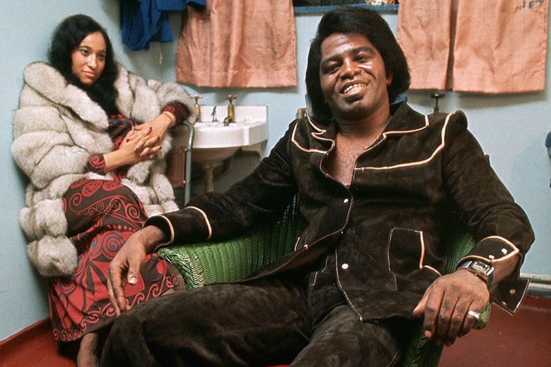 James Brown wears a brown suade suit and leans back in his armchair backstage, smiling to the camera.
