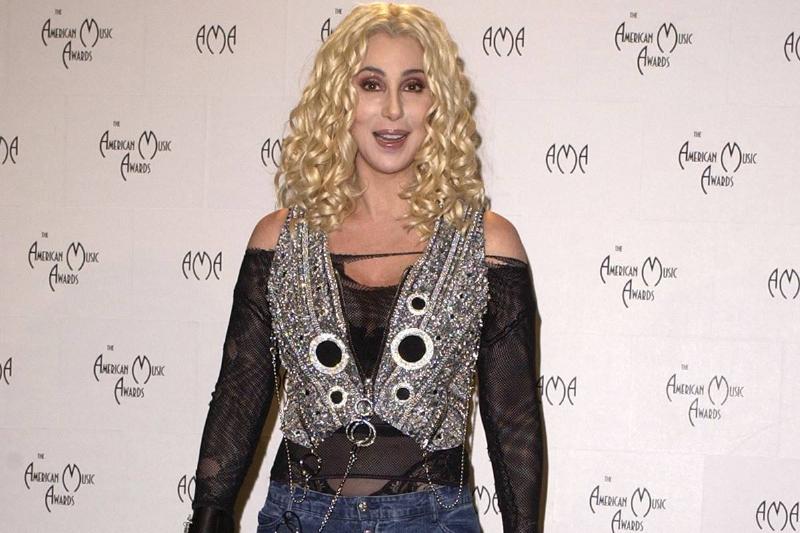 Cher has blonde, curled hair and wears a rock-inspired outfit.