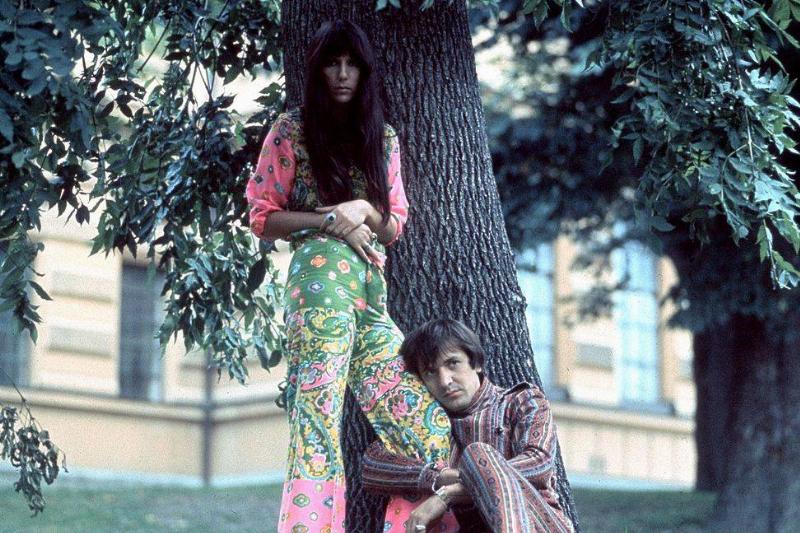 Sonny and Cher pose in pattered clothes in a park.